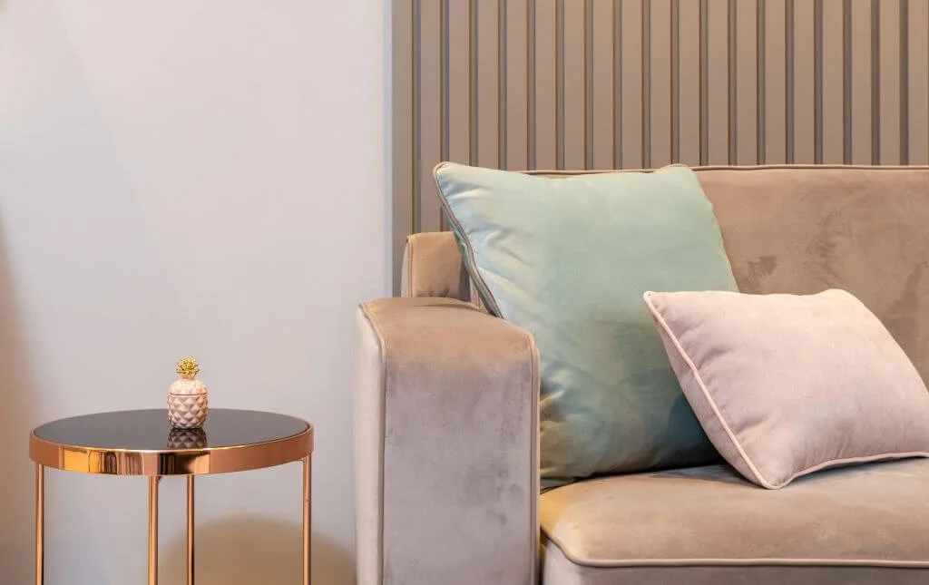 Šviesus, minimalistinis svetainės kampas su švelniai pilka sofa. Šviesios spalvos ir minimalus dekoras padeda mažam kambariui atrodyti erdvesniam.