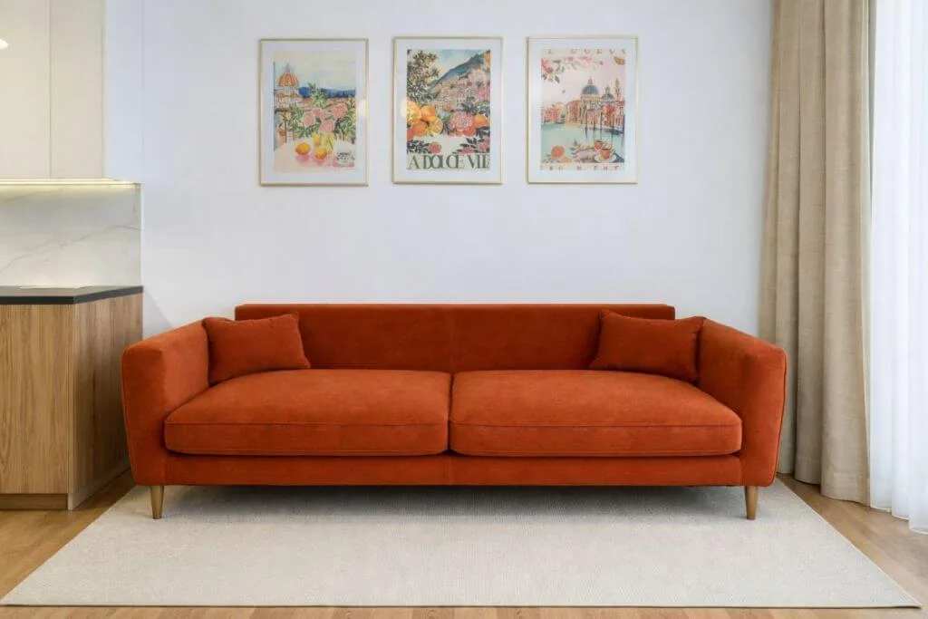 Moderni oranžinė sofa svetainėje su trimis paveikslais viršuje – idėja, kaip papuošti tuščią sieną virš sofos minimalistiniame interjere.
